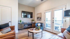 A spacious living room with a wooden floor, a sofa, a coffee table, a TV, and a balcony with a railing. - Chic Townhome | Walk to AT&T Stadium + Globe Life (Arlington)