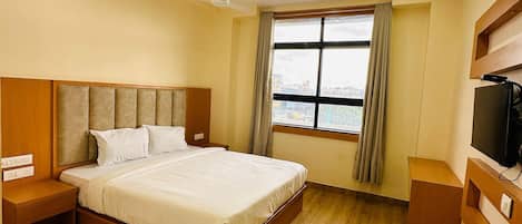 A hotel room with a wooden floor, a bed with white bedding, a ceiling fan, a window with curtains, and a TV on a wooden stand.