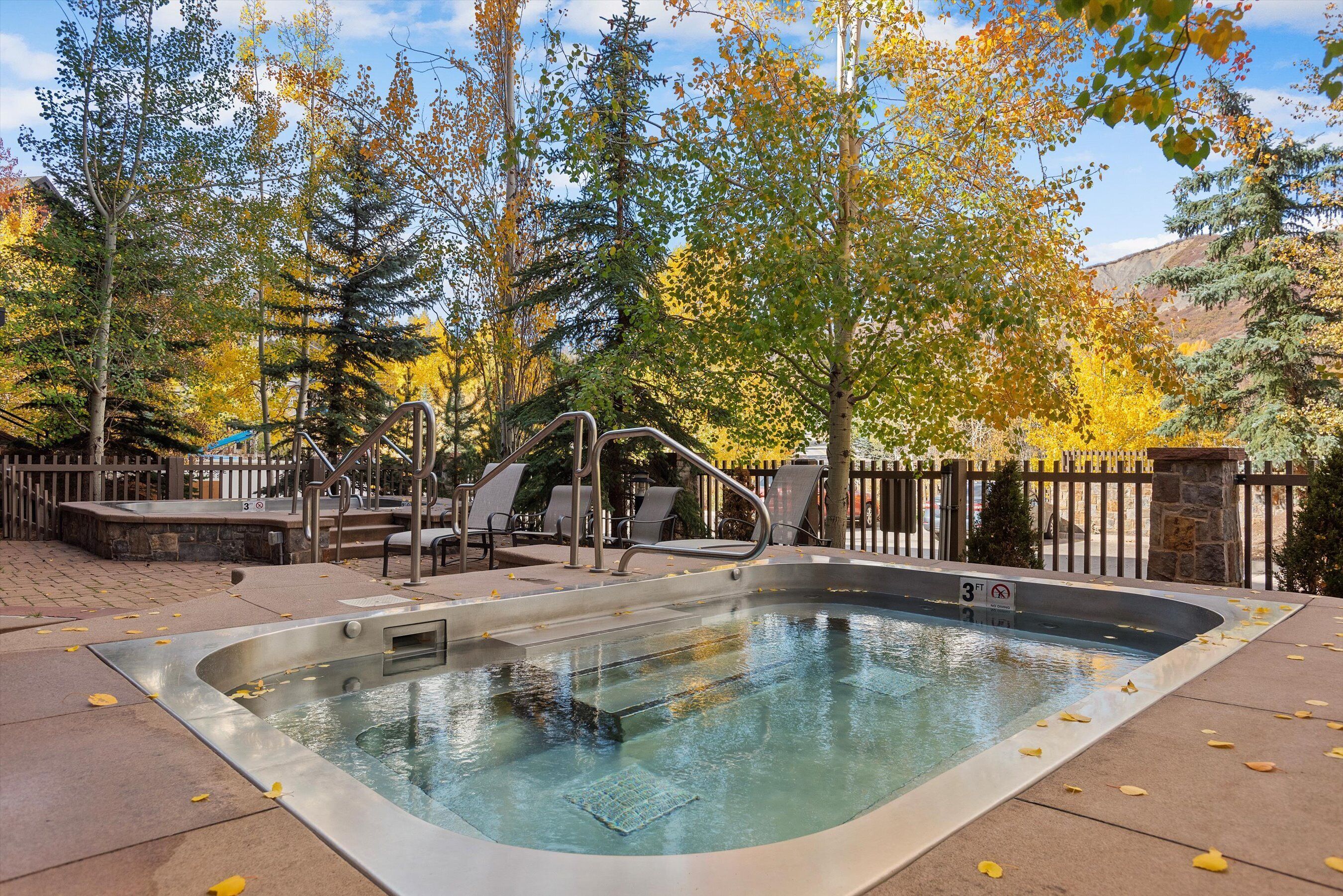 Un jacuzzi rodeado por una terraza de madera, con un muro de piedra y una valla metálica al fondo.