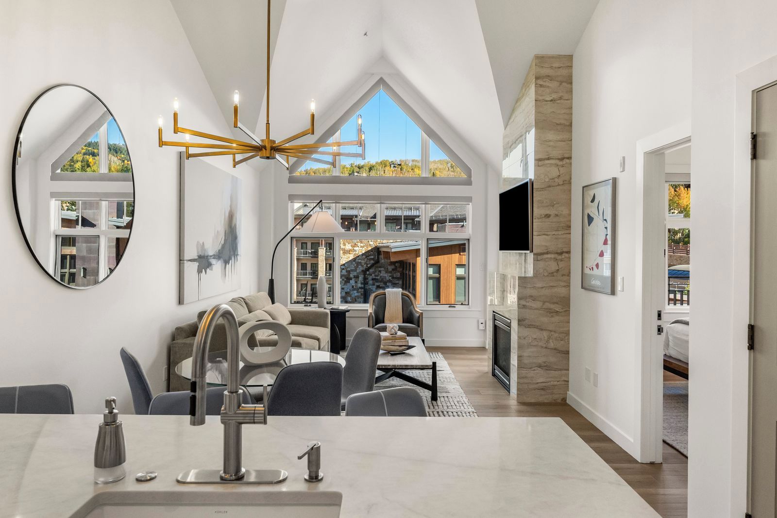 A modern living room with a high ceiling, a fireplace, a dining area with a kitchen counter, and a large window offering a view of a building and trees.