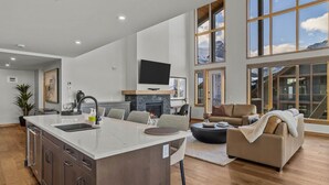 A spacious living area with a large island, a fireplace, and a view of mountains through large windows. - Mountain Therapy | Just Be Here (Canmore)