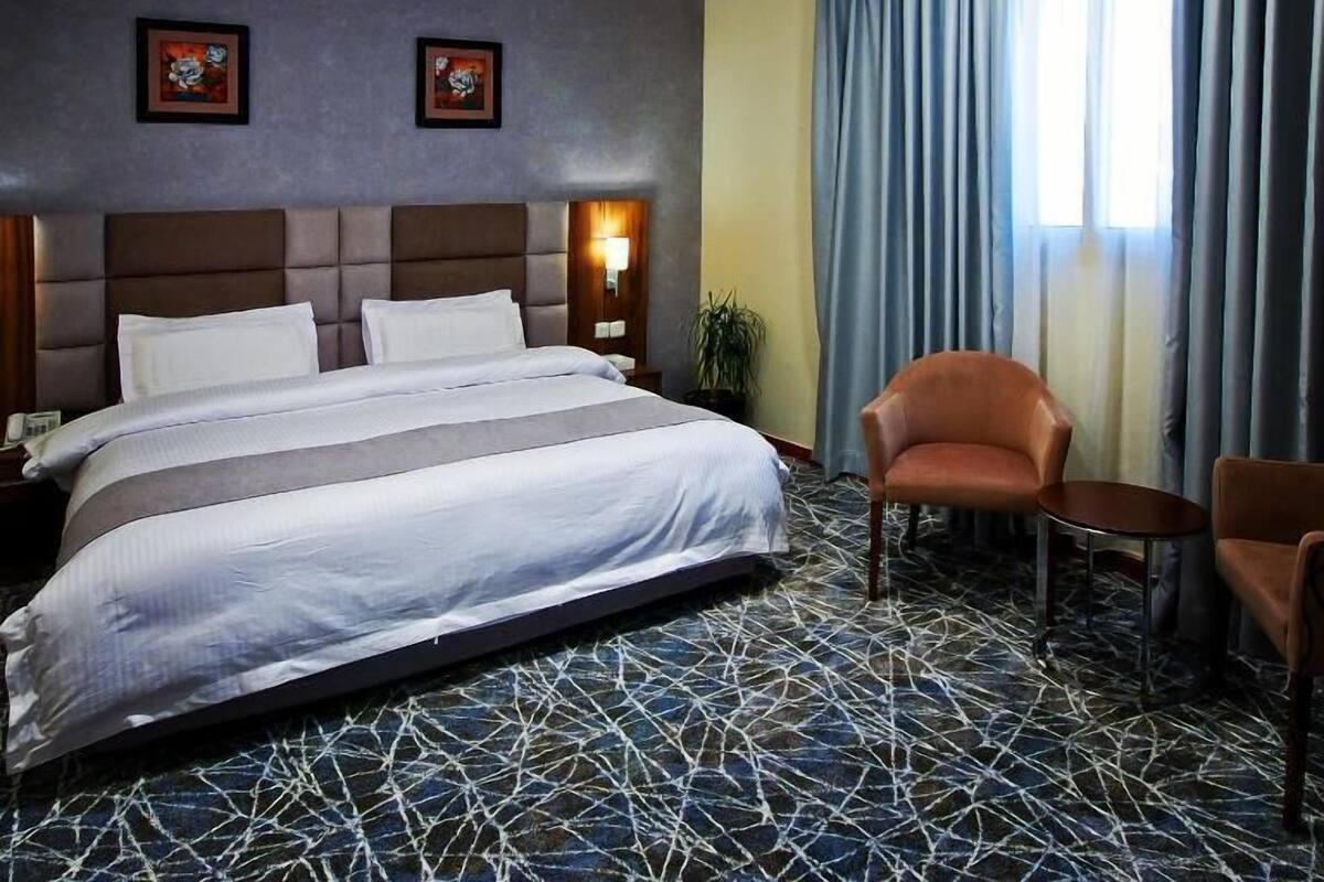 A hotel room with a large bed, two framed pictures on the wall, a chair, a small table, and a window with curtains.