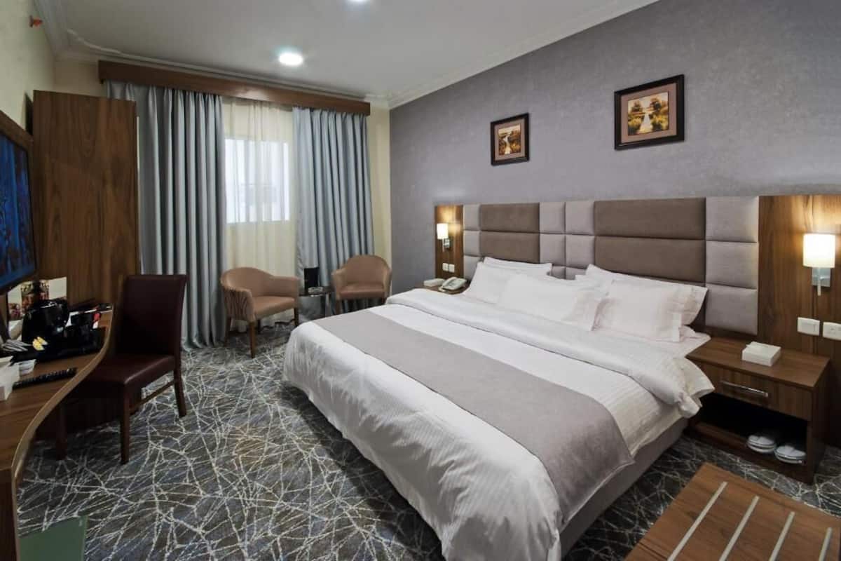 A hotel room with a large bed, two armchairs, a desk with a computer, and a window with curtains.