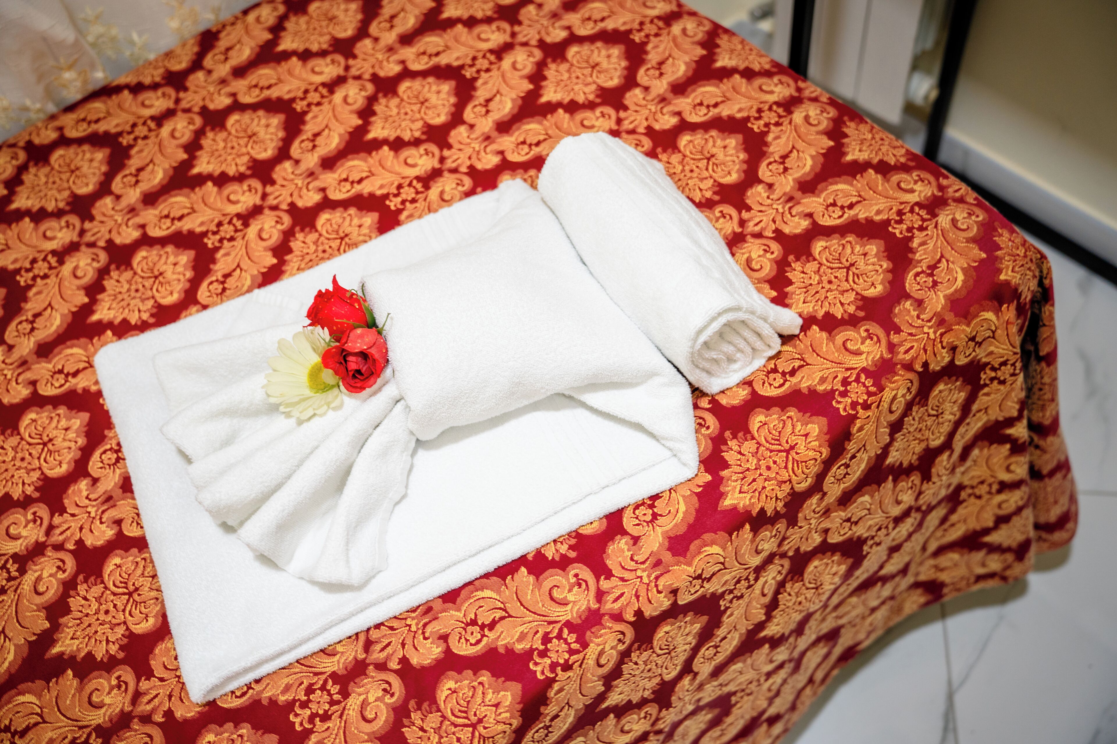 A neatly folded white towel with a red rose on a bed with a red and gold patterned bedspread.