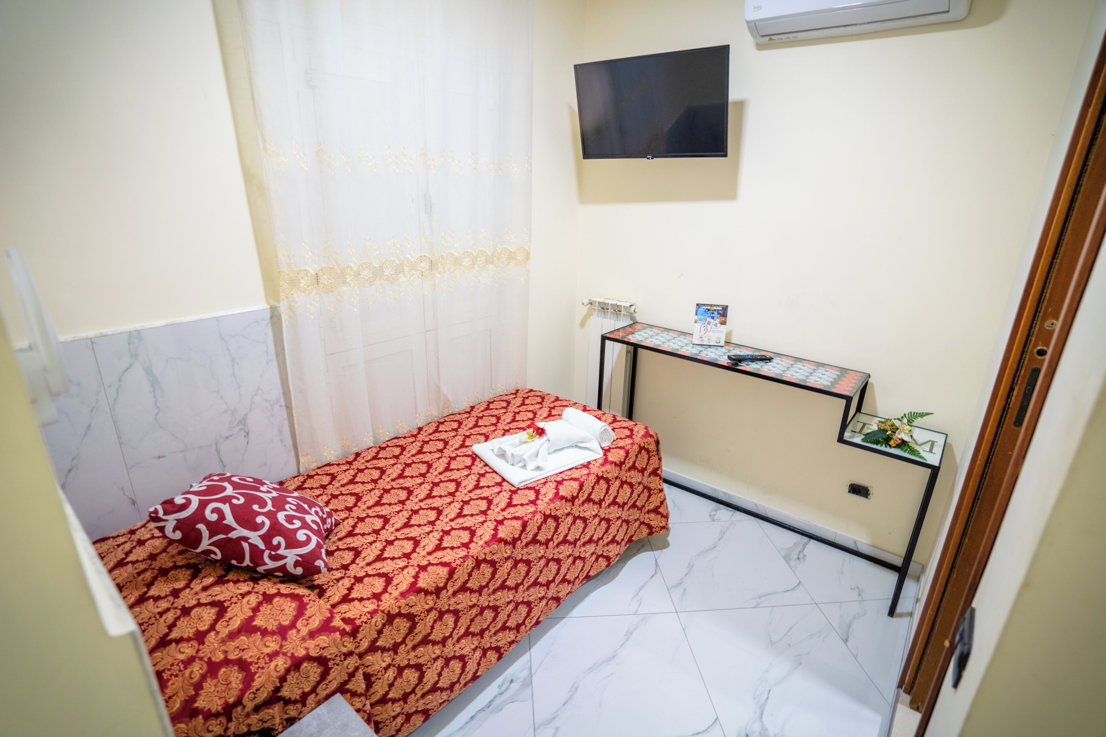 A small, neatly arranged room with a single bed, a bedside table with a lamp, and a television mounted on the wall.