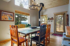 A dining area with a wooden table, chairs, and a chandelier. There are windows with blinds and a view of trees outside.