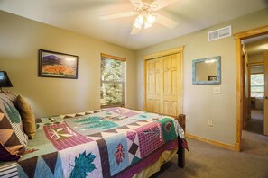 A bedroom with a large bed, a ceiling fan, a window with blinds, a door, and a framed picture on the wall.