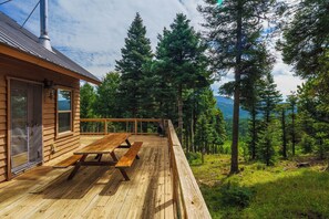 A wooden deck with picnic tables overlooking a forested area with tall trees and a mountain in the background.