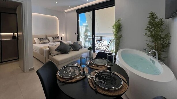 A modern, well-lit room with a dining table set for two, a sofa, and a large window offering a view of the city. - BlueMotion - PortSuite (Piraeus)