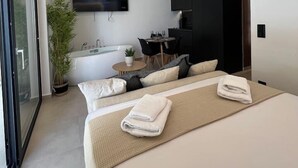 A modern hotel room with a large bed, a flat-screen TV mounted on the wall, a small round table with chairs, a potted plant, and a bathtub. - BlueMotion - PortSuite (Piraeus)