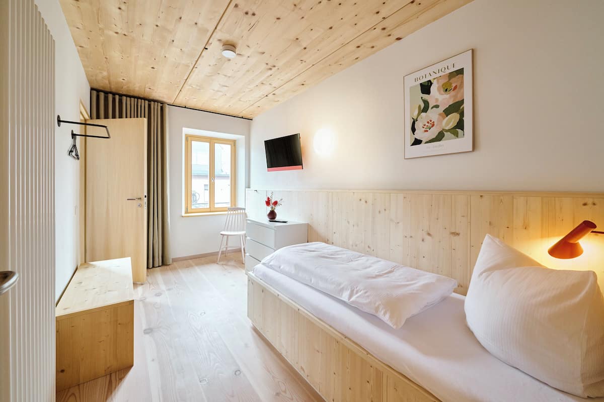 A bedroom with a wooden ceiling, a bed with white bedding, a desk with a chair, a TV mounted on the wall, and a window with curtains.