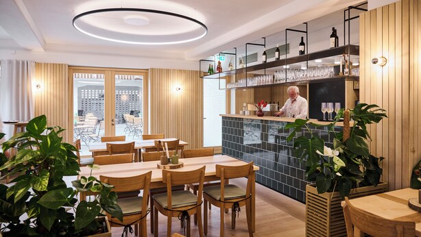 Un interior de restaurante moderno con mesas y sillas de madera, una zona de bar con estantes llenos de botellas y vasos, y plantas grandes en macetas.