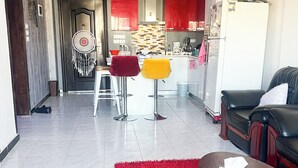 A modern living room with a red rug, a sofa, and a coffee table. There is a kitchen area with red cabinets and a white refrigerator.