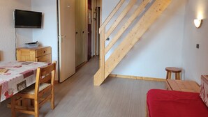 A room with a wooden staircase, a television mounted on the wall, a dining table with chairs, and a red bench. - Cozy apartment near slopes, valley view (La Plagne Montchavin Les Coches)