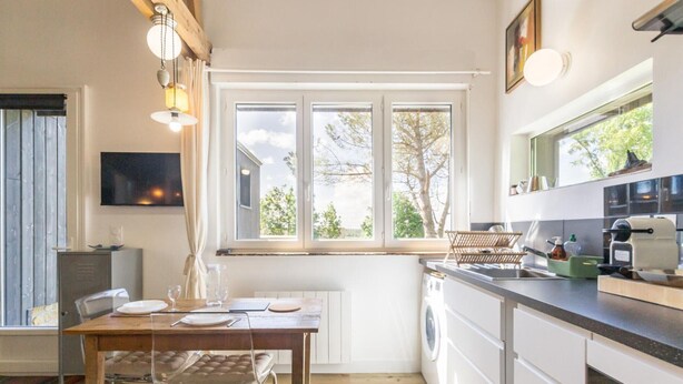 A modern kitchen with a wooden table, a chair, a washing machine, and a window with a view of trees.