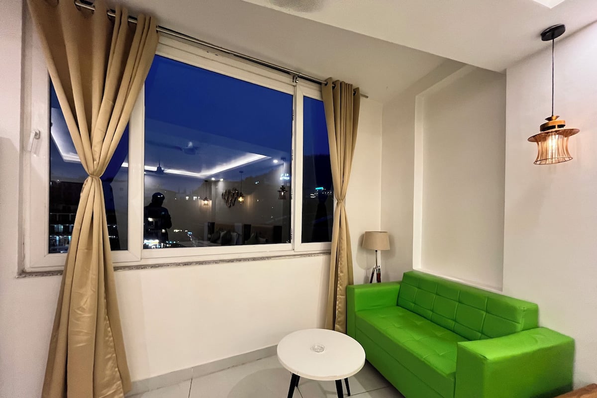 A room with a green sofa, a small white table, a lamp, and a window with curtains.