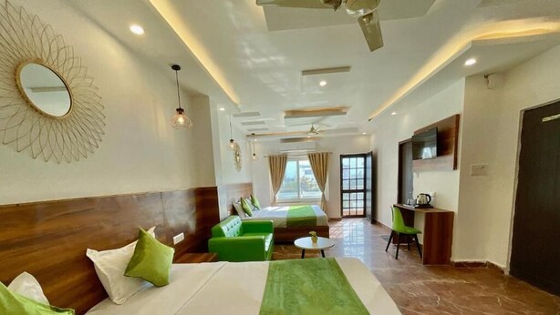 A hotel room with a large bed, green and white bedding, a green sofa, a small round table, a TV, and a ceiling fan. - Wanna Stay Rishikesh (Rishikesh)