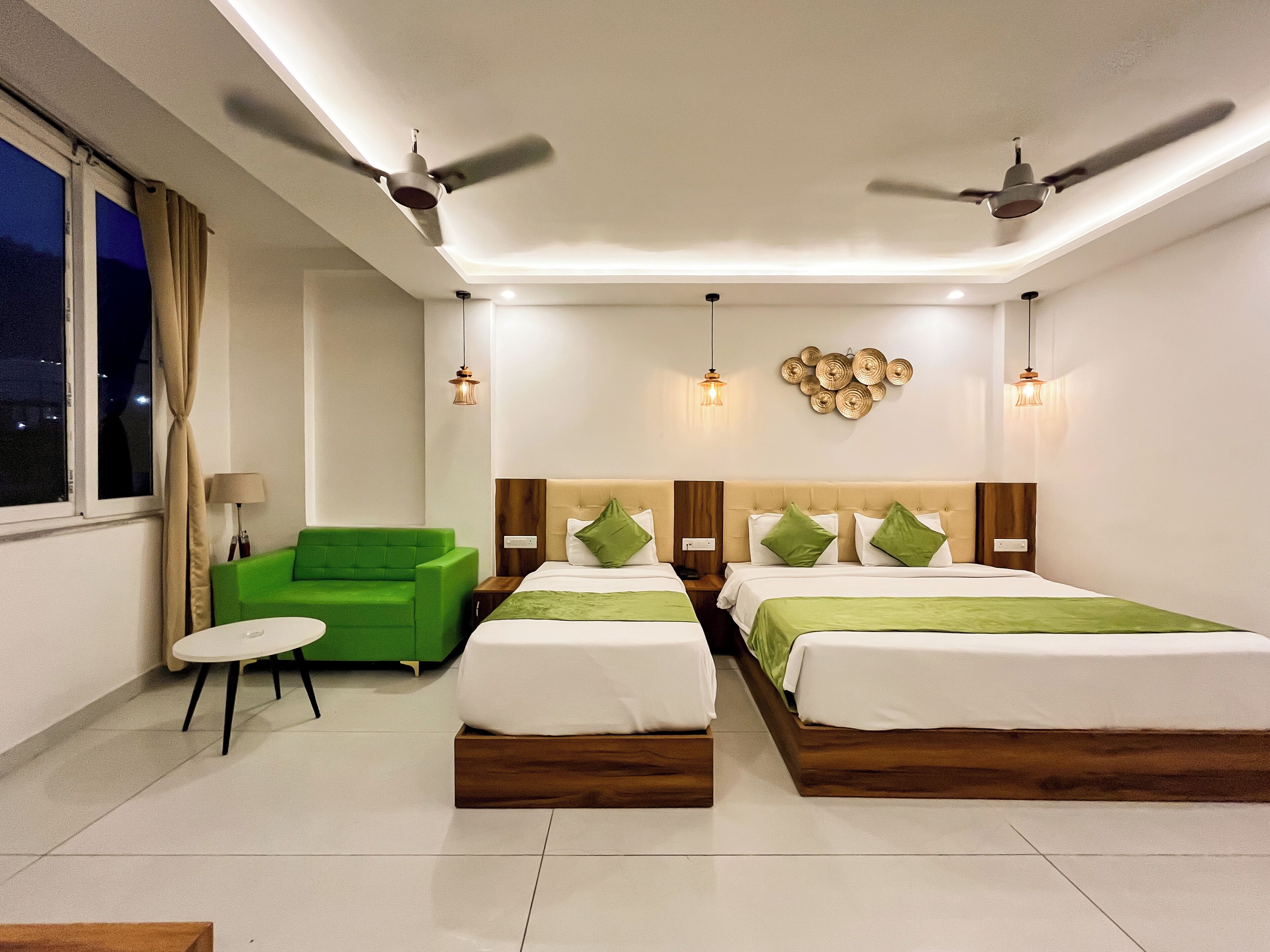 A hotel room with two beds, a green armchair, a small table, and ceiling fans.