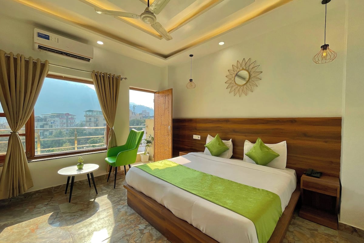 A hotel room with a large bed, a green chair, a small table, and a view of the city through the window.