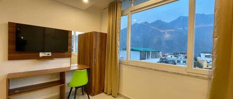 A room with a TV mounted on the wall, a desk with a chair, a toilet, and a large window with a mountain view.