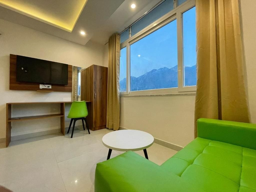 A modern room with a green sofa, a small white table, a green chair, a TV mounted on the wall, and a window with a mountain view.