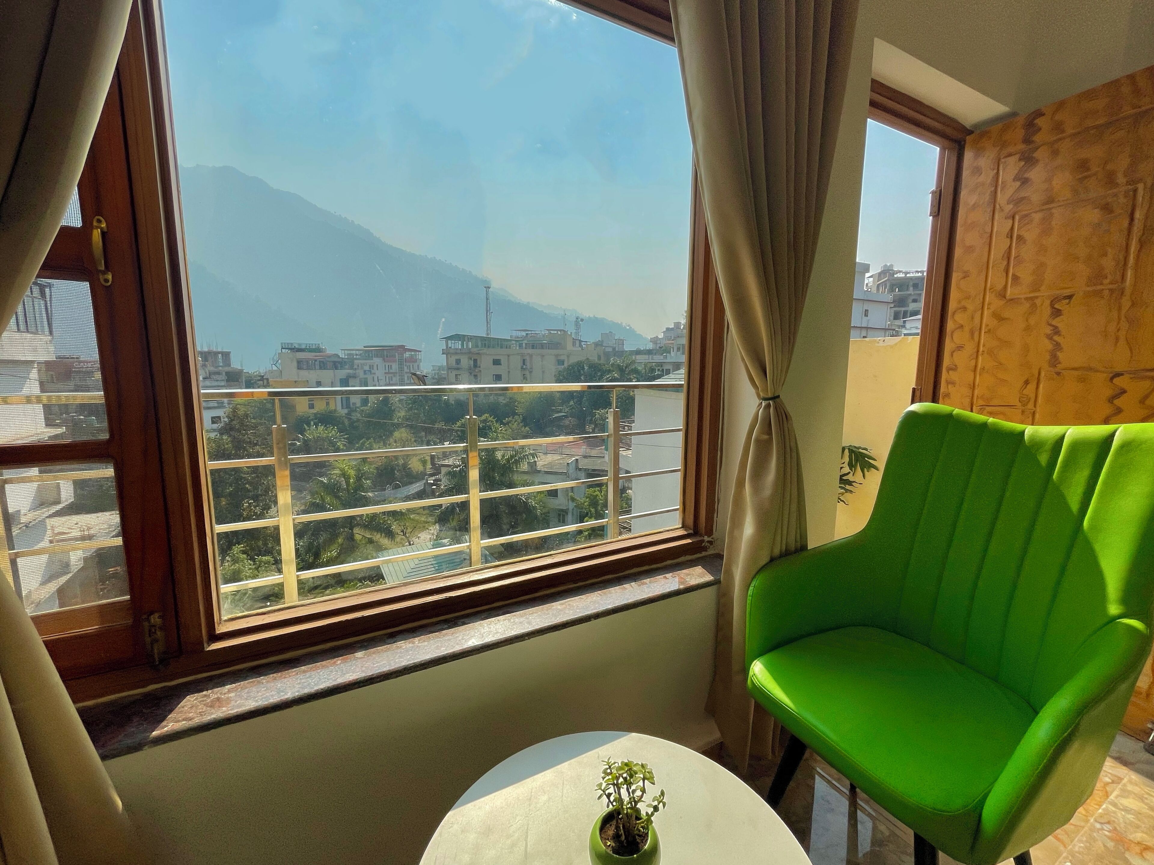 A room with a green chair, a small table with a plant, and a view of buildings and mountains through large windows.