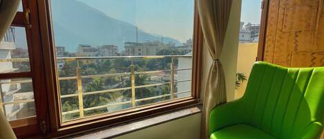 A room with a green chair, a small table with a plant, and a view of buildings and mountains through large windows.