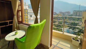 A room with a green chair, a small table with a plant, a window with curtains, and a view of buildings and mountains. - Wanna Stay Rishikesh (Rishikesh)