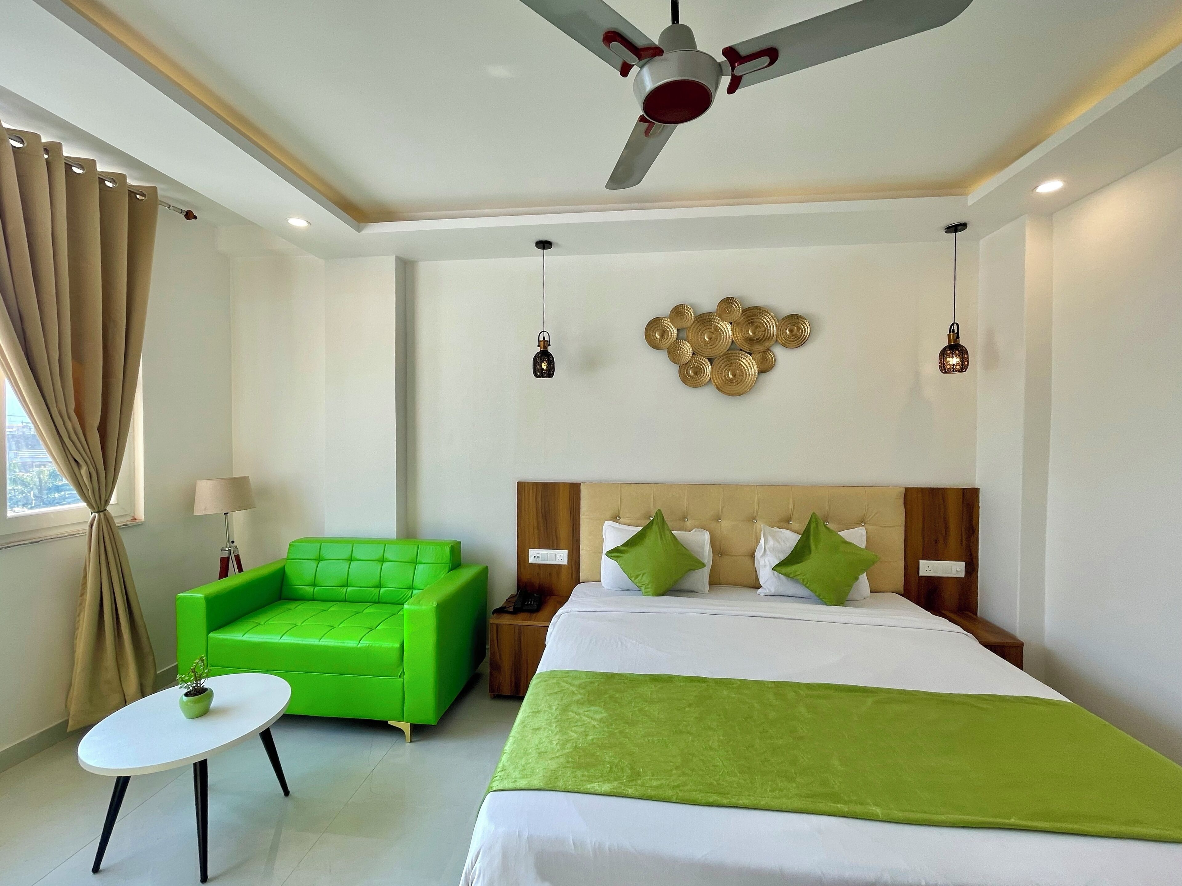 Une chambre d’hôtel avec un lit, un fauteuil vert, une petite table, un ventilateur de plafond et un élément décoratif mural.