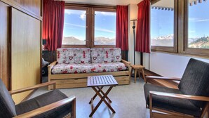 Ein gemütliches Zimmer mit einer Couch, zwei Sesseln, einem kleinen Tisch und großen Fenstern mit Blick auf die Berge.
