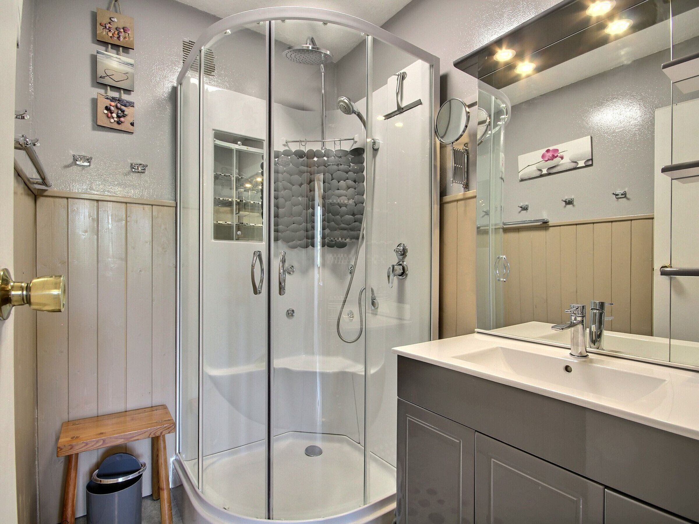 A modern bathroom with a glass-enclosed shower, a vanity with a mirror, and a small wooden stool.