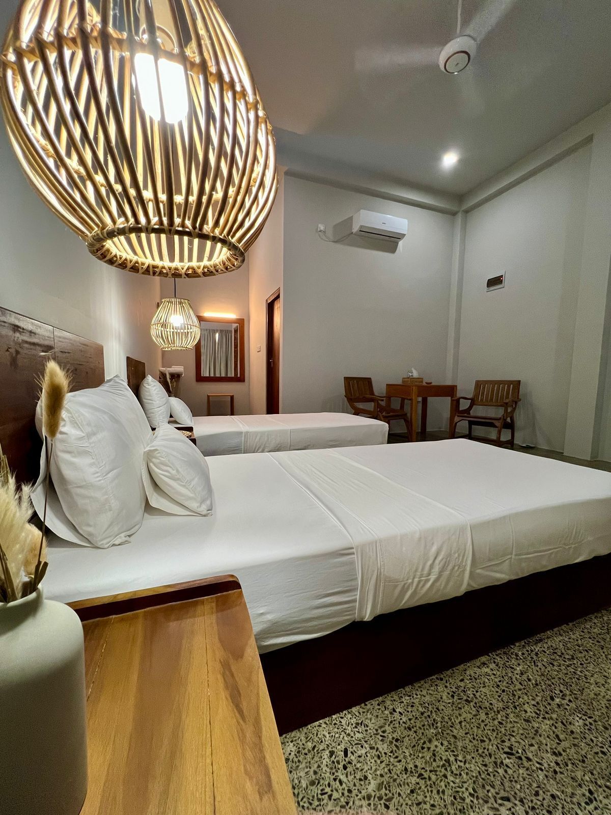 A hotel room with two beds, a wooden headboard, a woven light fixture, a small table, and a chair.