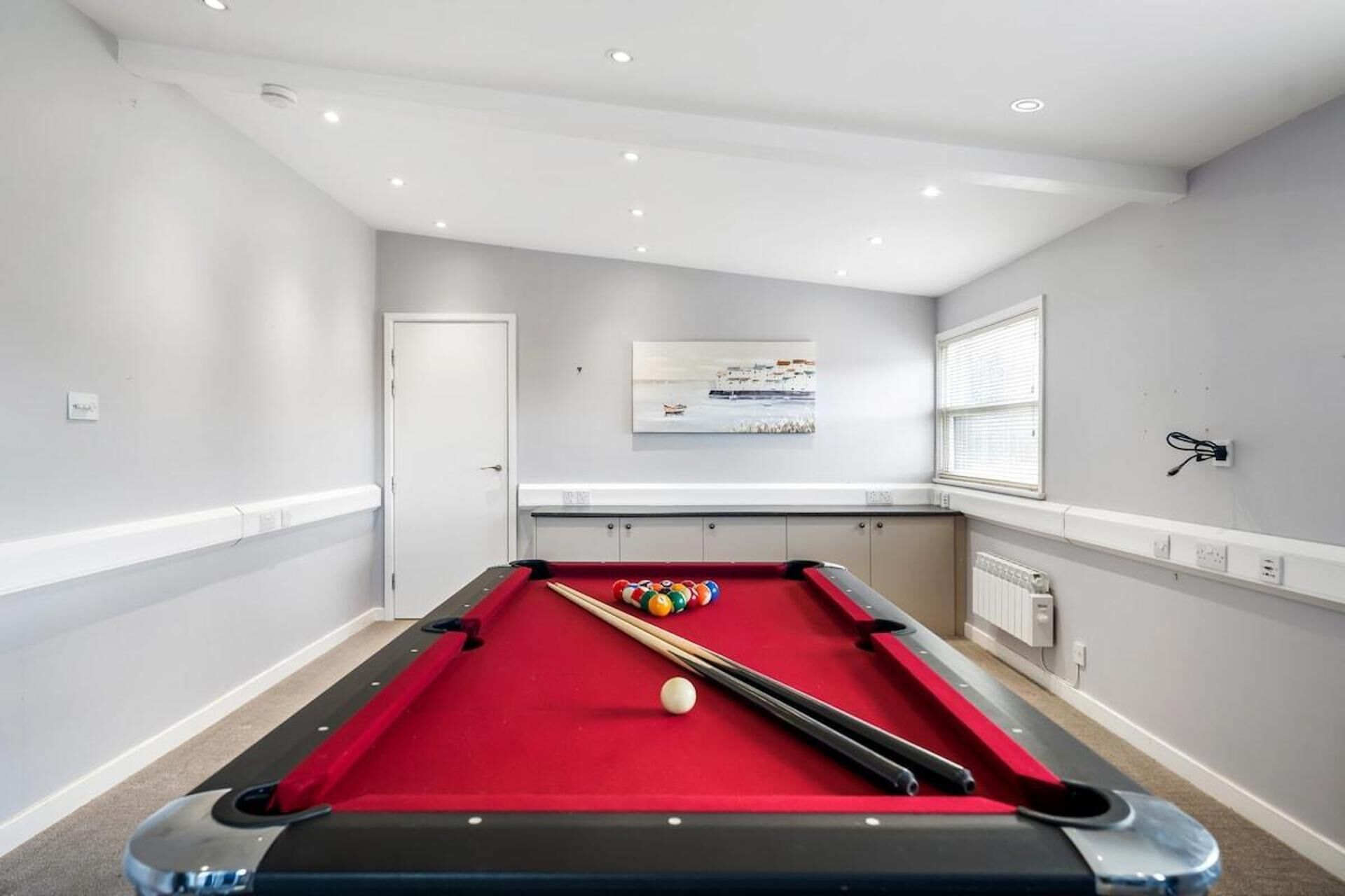 A pool table with balls, a door, a window with blinds, and a painting on the wall.