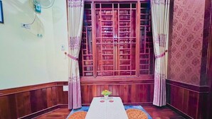 A room with a wooden floor, a table with a white tablecloth, a blue furry rug, and a window with wooden shutters. - Canary Homestay (Hue)