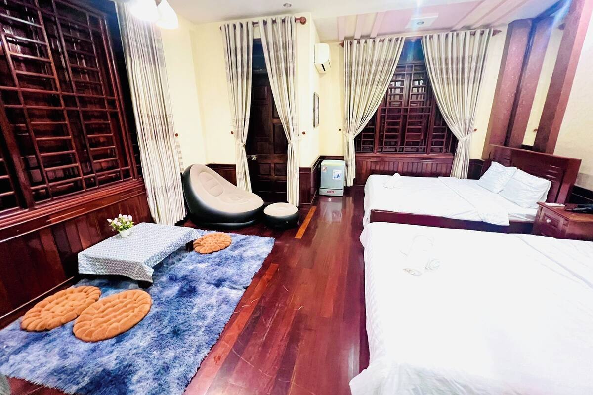 Quarto de hotel com duas camas, uma cadeira de massagem, uma mesinha com uma planta e um tapete azul com padrões circulares.