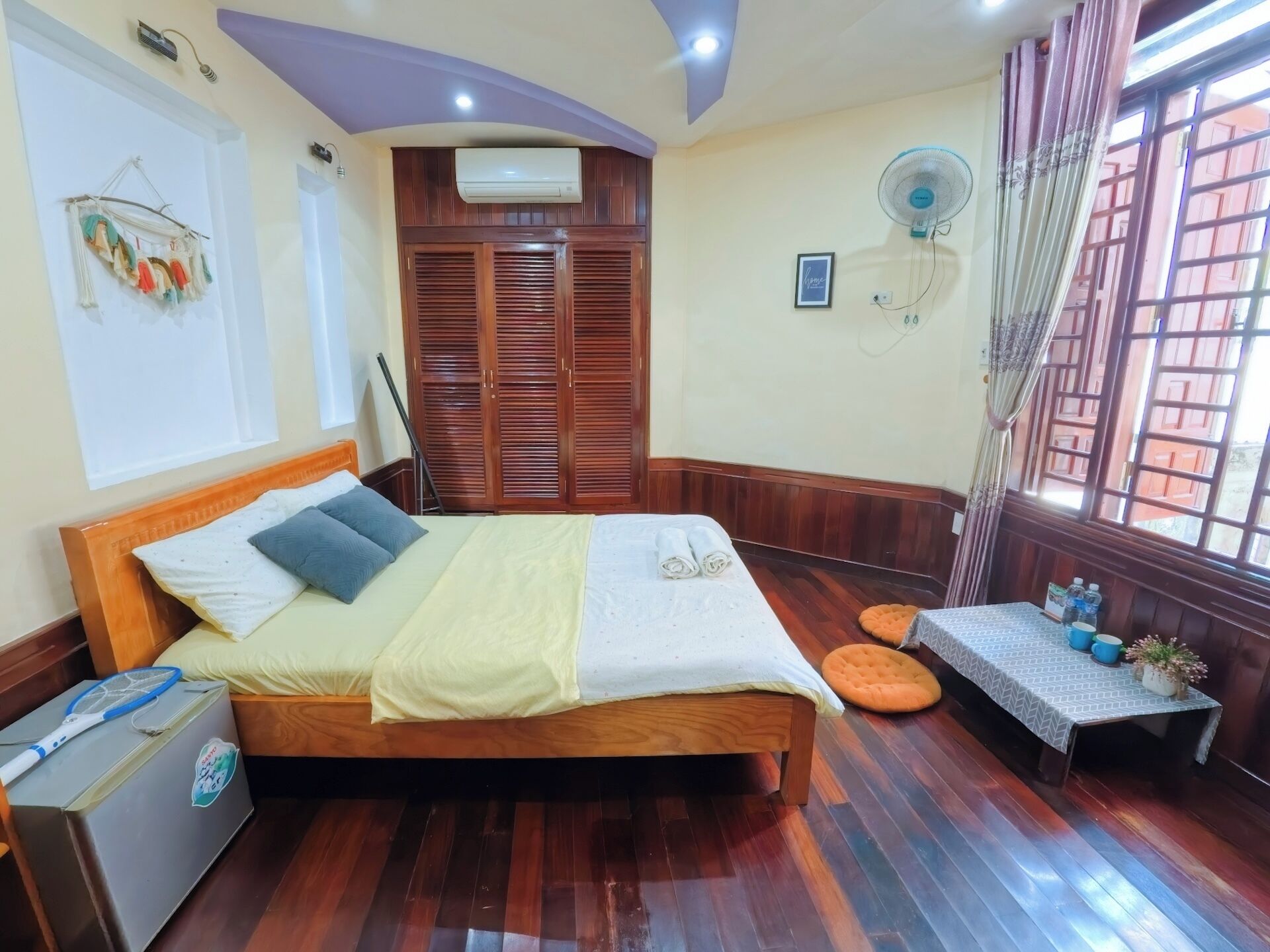A bedroom with a wooden bed, a bedside table, a small table with a chair, a window with curtains, and a wall-mounted fan.