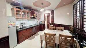 A kitchen with wooden cabinets, a refrigerator, a dining table with chairs, and a window with bars. - Canary Homestay (Hue)