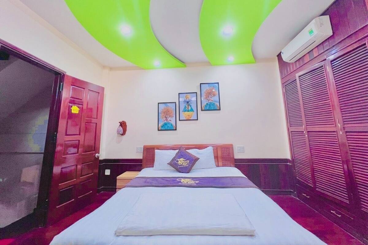 Quarto de hotel com uma cama, uma porta, parede com três quadros e teto com iluminação verde.