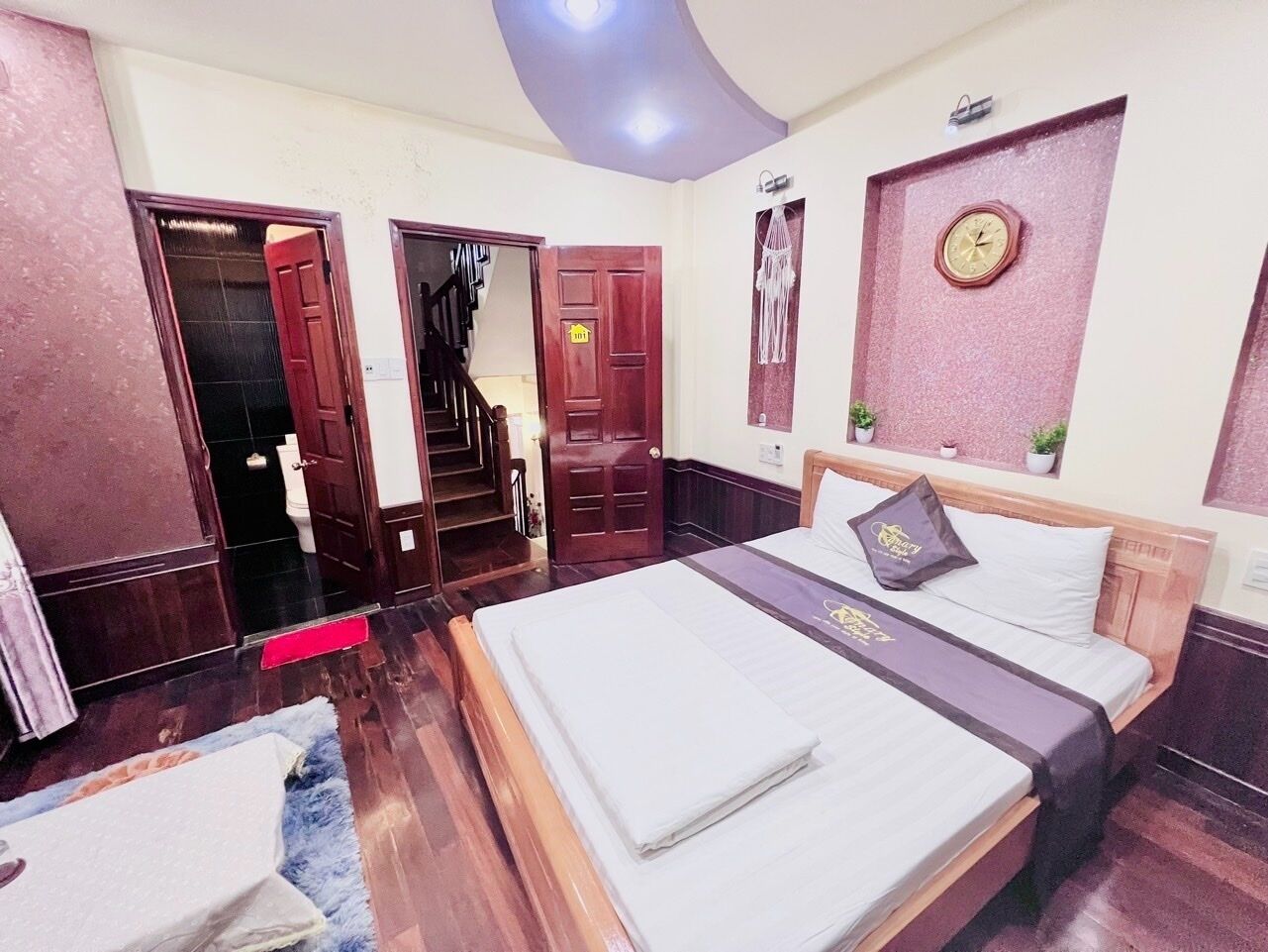 A hotel room with a large bed, a wooden wardrobe, a small desk, and a bathroom with a toilet and sink.
