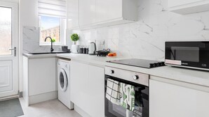 A modern kitchen with white cabinetry, a built-in washing machine, a microwave, and a window with blinds. - 3br-sleeps8-parking-wifi-closestation (Corby)