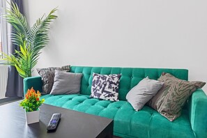 A green sofa with patterned cushions, a remote control, and a potted plant with orange flowers. - 3br-sleeps8-parking-wifi-closestation (Corby)