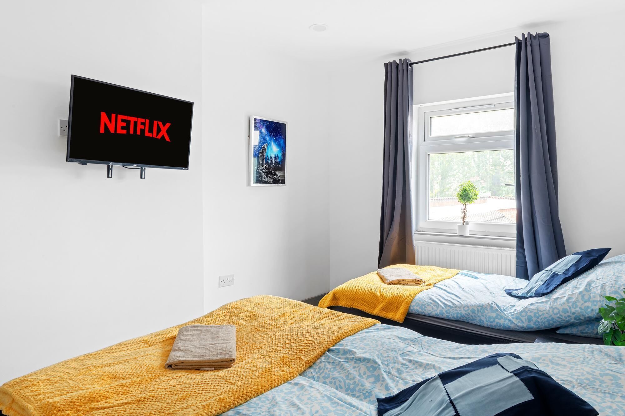 Quarto com duas camas, uma televisão exibindo o logotipo da Netflix, um quadro e uma janela com cortinas.