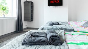 A hotel room with two beds, a TV mounted on the wall displaying 'NETFLIX', a radiator, a window with curtains, and a plant outside. - 3br-sleeps8-parking-wifi-closestation (Corby)