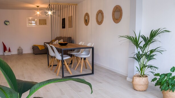 A modern living room with a dining area, featuring a light-colored herringbone floor, wicker furniture, and several potted plants. - Apartamento con terraza y piscina comunitaria en Puerto de la Cruz · 4 pax (Puerto de la Cruz)