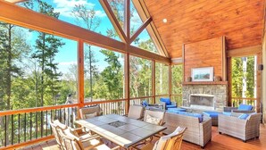 Una terraza de madera con chimenea de piedra, asientos al aire libre y grandes ventanales con vistas a un bosque.