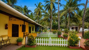 A yellow building with a white picket fence, surrounded by lush greenery and palm trees. - Vinsons Cottages (Colva)