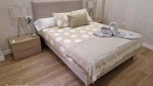 A neatly made bed with a polka dot bedspread, two pillows, and a towel on the side. There is a bedside table with a lamp and a clock. The room has wooden flooring and light-colored walls.