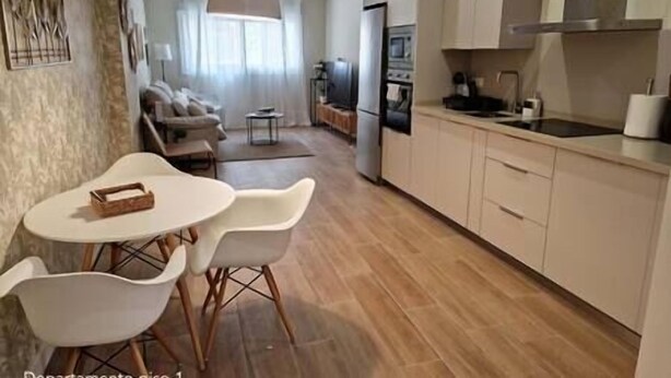 A modern kitchen with white cabinets, a dining area with a table and chairs, and a living room with a sofa and TV.