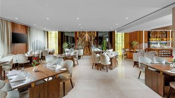 A modern hotel dining area with multiple tables, chairs, a flat-screen TV, and a decorative wall.