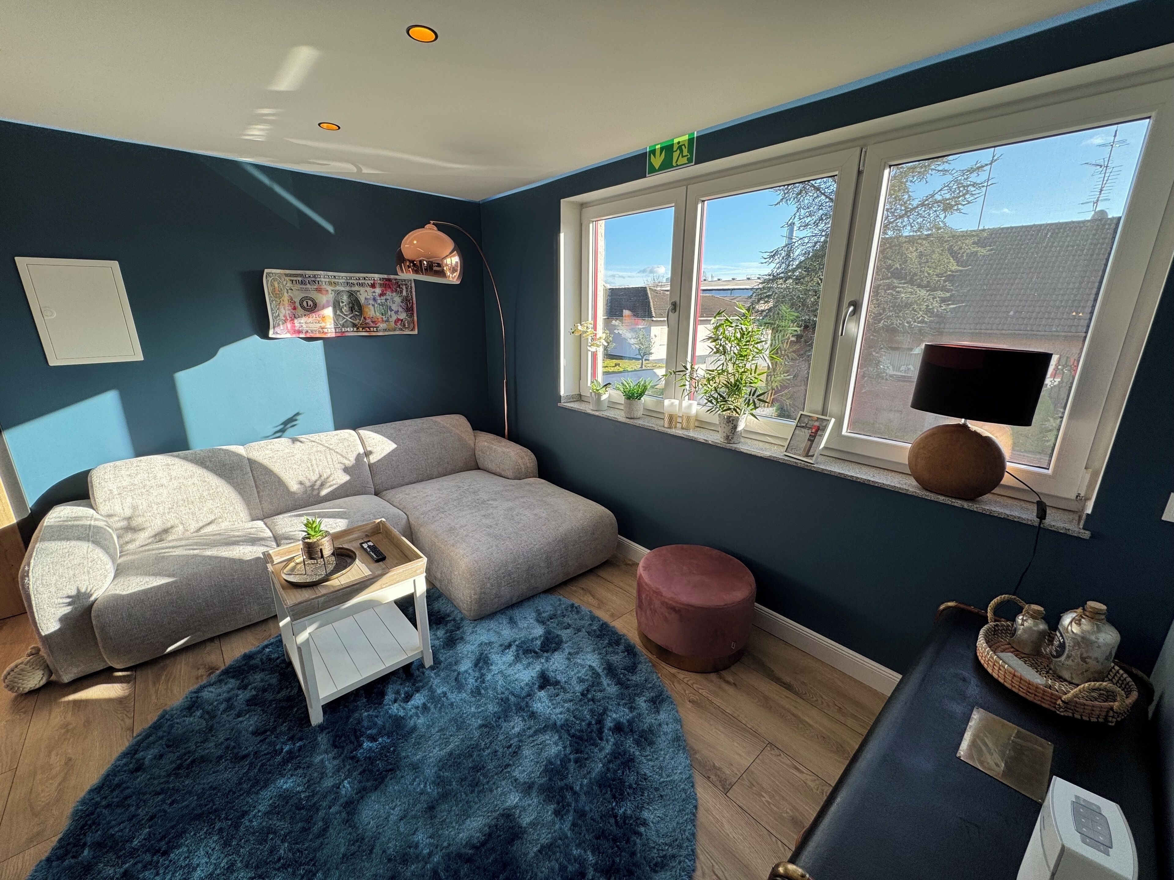 Ein gemütliches Wohnzimmer mit einem Ecksofa, einem flauschigen blauen Teppich, einem kleinen weißen Tisch und einem Fenster mit Blick nach draußen.